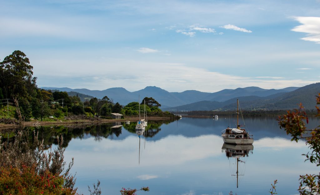A calm lake with sailboats anchored on still water, reflecting the surrounding trees, hills, and distant mountains under a partly cloudy sky.
