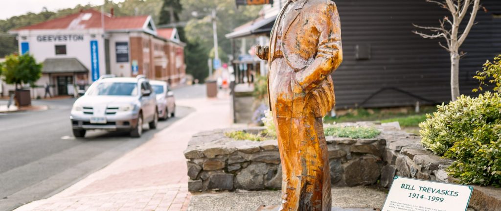 A wooden statue of a man in a suit stands on a stone platform by a sidewalk; a sign reads Bill Trevaskis 1914–1999. Cars and buildings are visible along the street in the background.