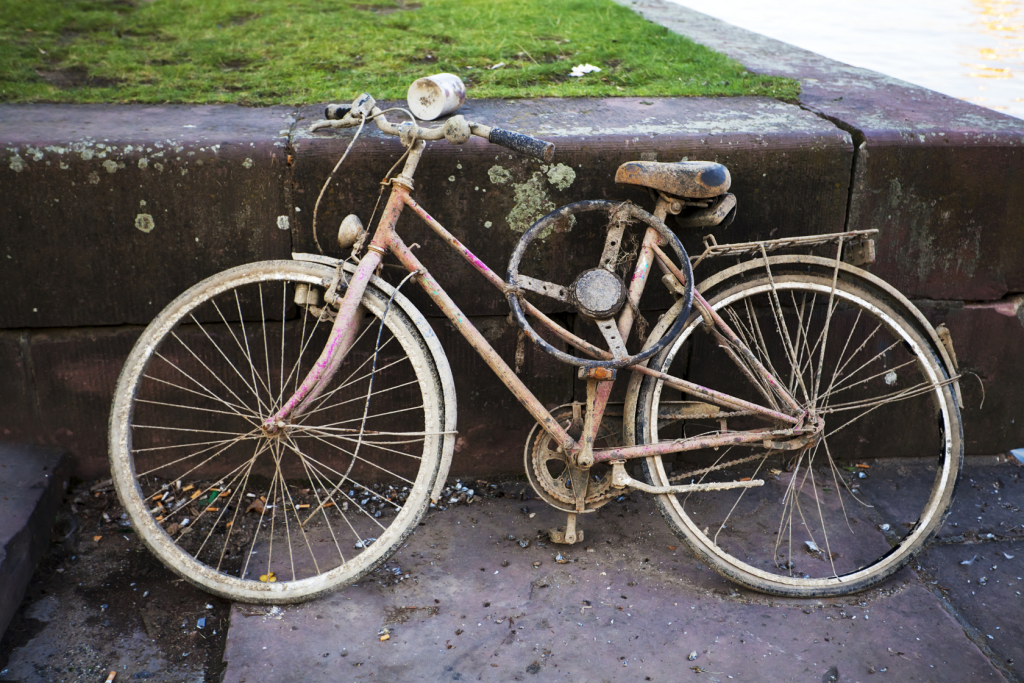 A rusty, mud-covered bicycle with worn tires is leaning against a stone wall near a grassy area and a body of water. The bike appears old and abandoned.