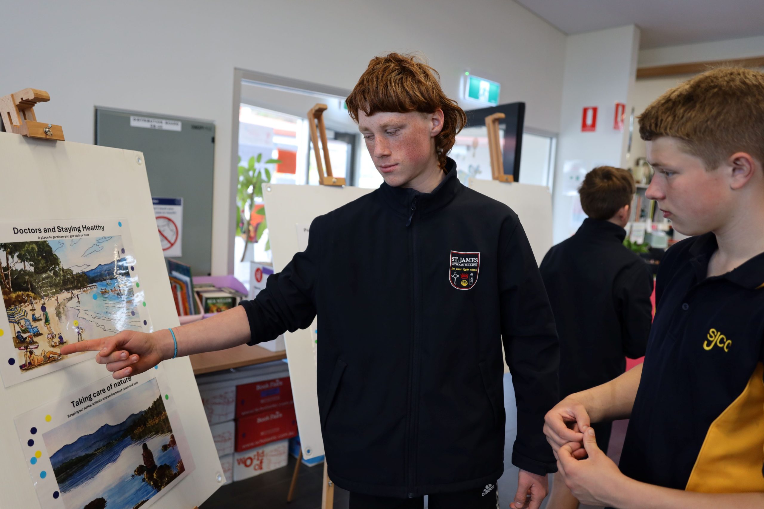 Two students stand indoors by a display board. One student points at images and text on the board, while the other listens attentively. The environment looks like a classroom or school exhibition.