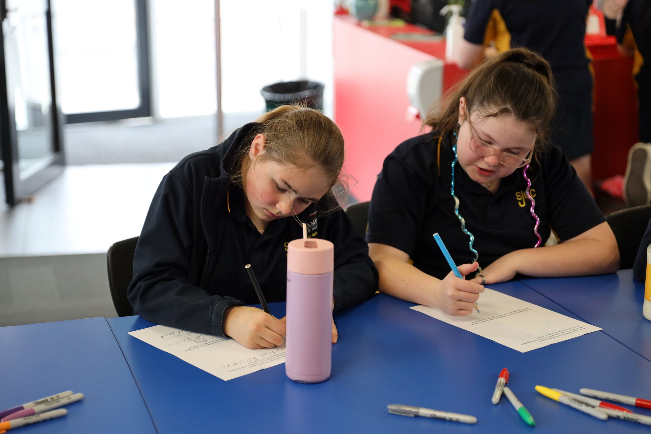 Two girls sit at a blue table writing on paper with pens. There are colorful markers and a pink water bottle on the table. Both are focused on their work in a bright indoor setting.