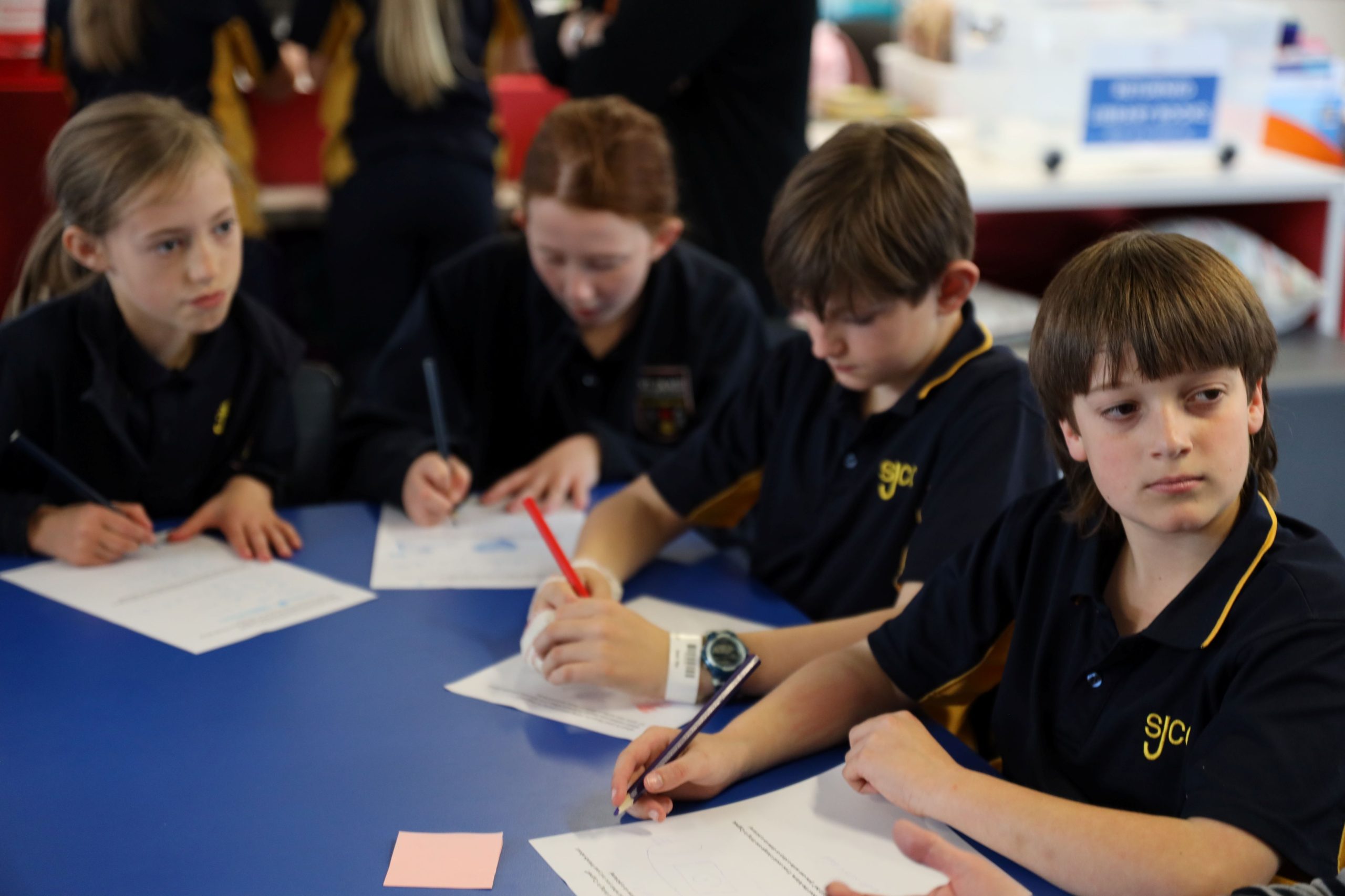 Four school children in dark uniforms sit around a blue table, writing on sheets of paper with pens. They appear focused and engaged in their activity in a classroom setting.