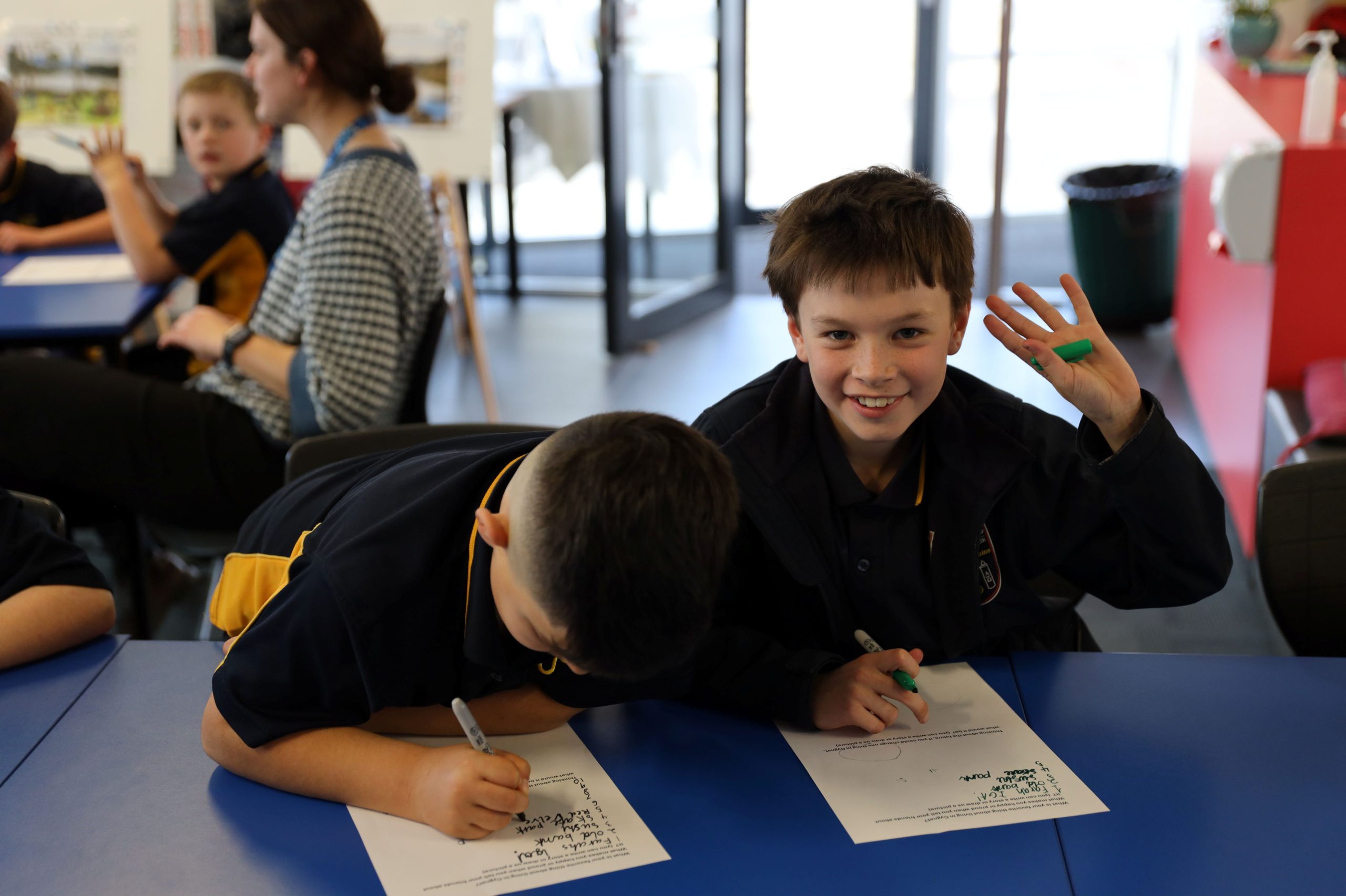 Two boys sit at a blue table in a classroom. One boy writes on a paper while the other smiles at the camera and holds up four fingers. Other students and an adult are visible in the background.