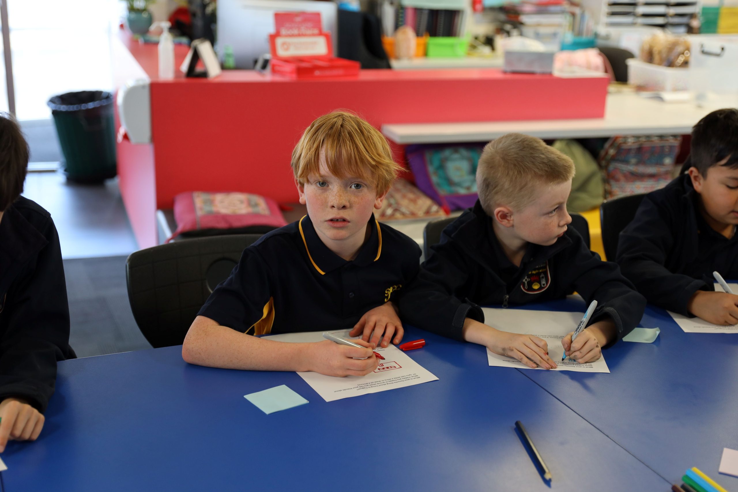 Three young boys sit at a blue table in a classroom, working on worksheets with pens. One boy with red hair looks at the camera while the others focus on their papers. Classroom supplies and bags are visible in the background.