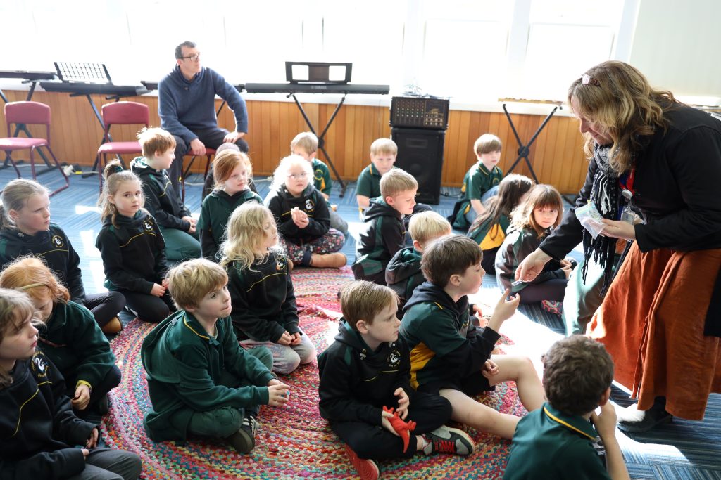 A group of young children sit cross-legged on a colorful rug in a classroom, attentively facing a woman who is interacting with one child. An adult sits in the background near musical instruments and windows.