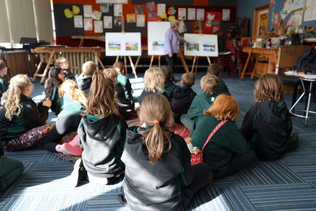A group of children sit on a classroom floor, facing an adult who is presenting pictures on easels. The classroom walls are decorated with colorful artwork and posters.