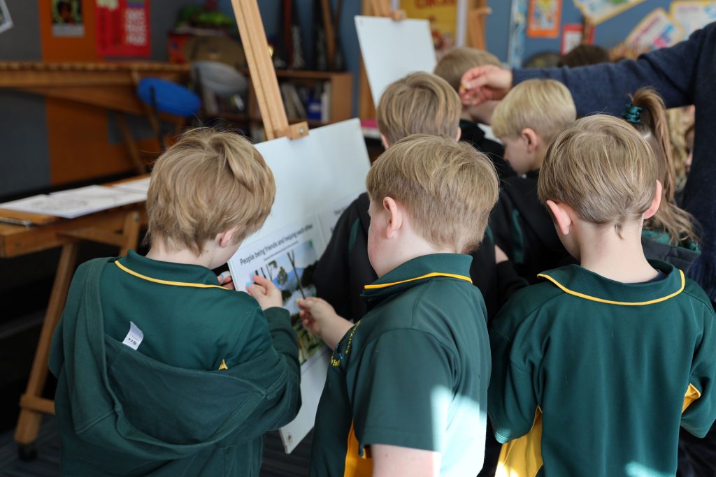 A group of young children in green uniforms stand closely together, looking at and interacting with a display board in a classroom setting. An adult hand is visible, assisting them.