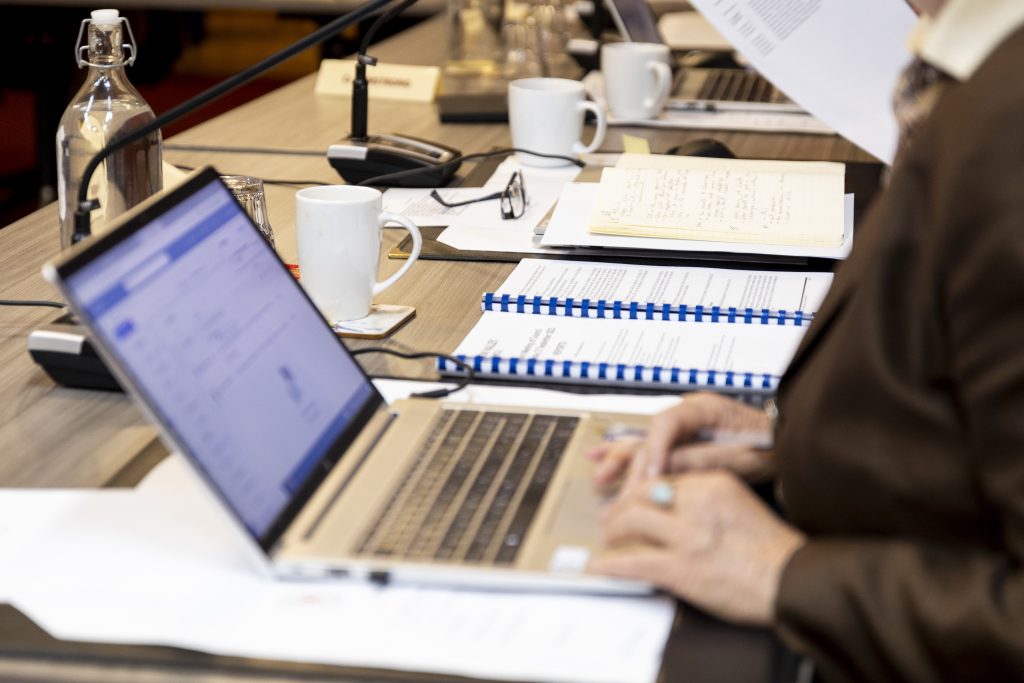 A person is typing on a laptop at a conference table, surrounded by documents, notebooks, coffee mugs, a glass water bottle, and microphones. Other people are seated nearby, engaging in paperwork.