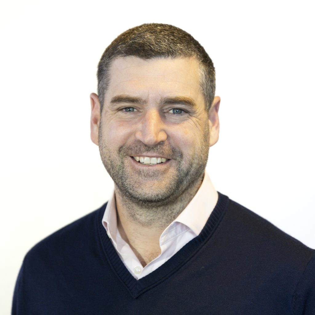 A man with short dark hair and a beard is smiling, wearing a navy blue sweater over a light pink collared shirt, against a plain white background.