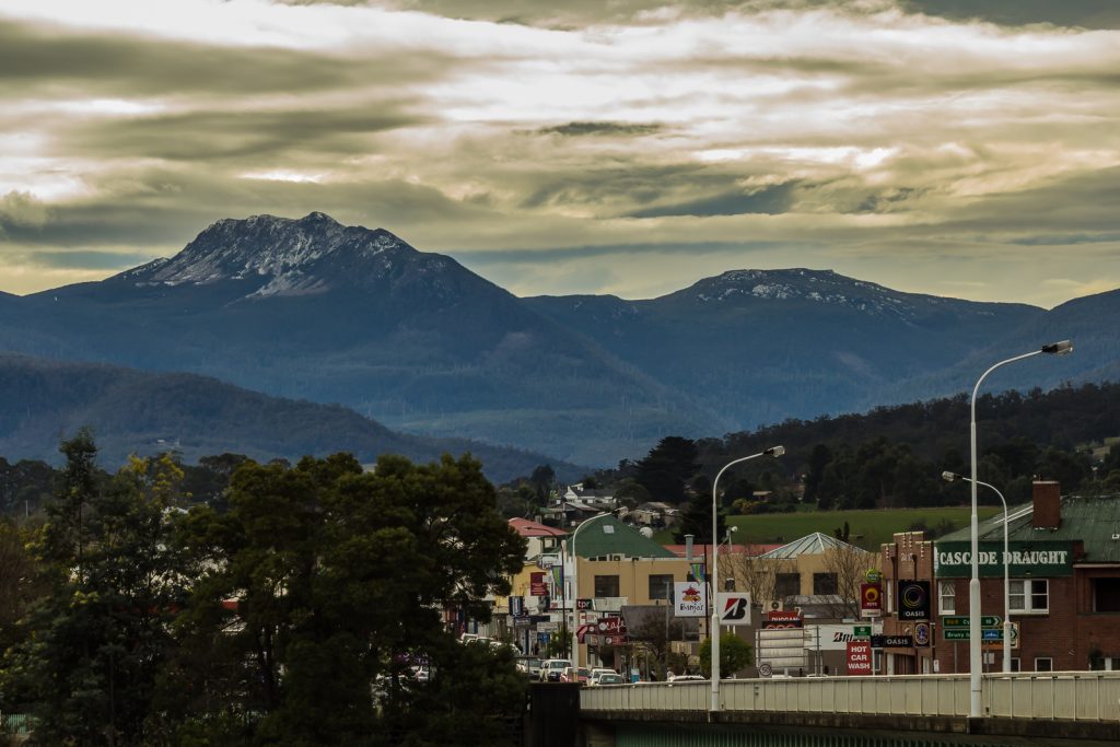 A small town with shops and streetlights is set against a backdrop of forested hills and tall, rugged mountains under a cloudy sky.