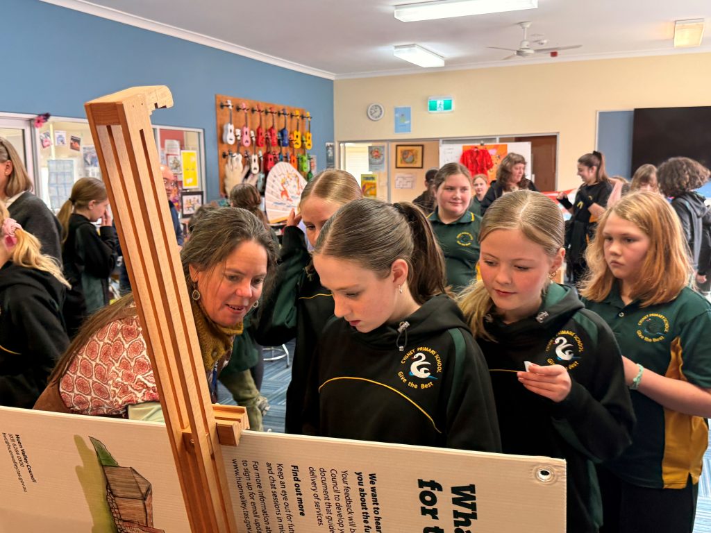A group of schoolgirls in matching uniforms gathers around an exhibit with a female teacher, reading and discussing the display in a colorful classroom.