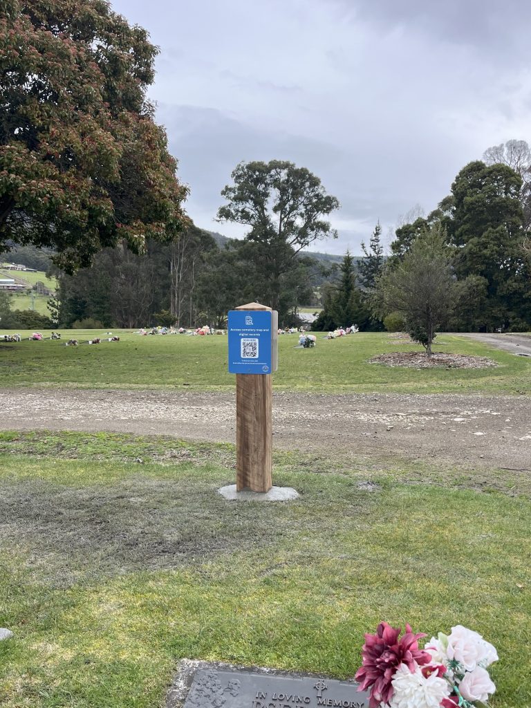 A wooden post with a blue sign and QR code stands on grassy ground in a cemetery, with headstones, trees, and hills visible in the background under a cloudy sky. Pink and white flowers are in the foreground.