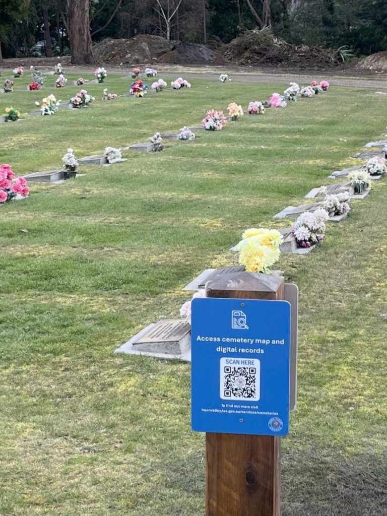 A blue sign with a QR code for accessing the cemetery map and digital records stands in front of rows of graves adorned with flower arrangements in a grassy cemetery.