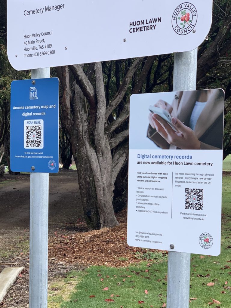 Three signs at Huon Lawn Cemetery: one large sign with contact info, and two smaller signs about accessing digital cemetery records via QR code, set in front of trees.