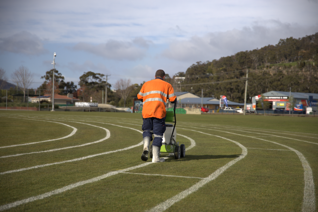A worker in an orange high-visibility jacket and white boots is painting white lines on a grass sports field using a line marking machine. The scene is outdoors with buildings and hills in the background.