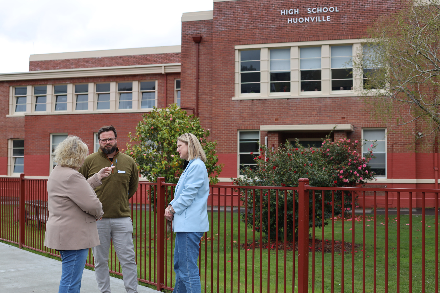 Three adults stand talking outside a red-brick school building with High School Huonville sign. Two women and one man are near a red fence, with green lawns and blooming bushes behind them.