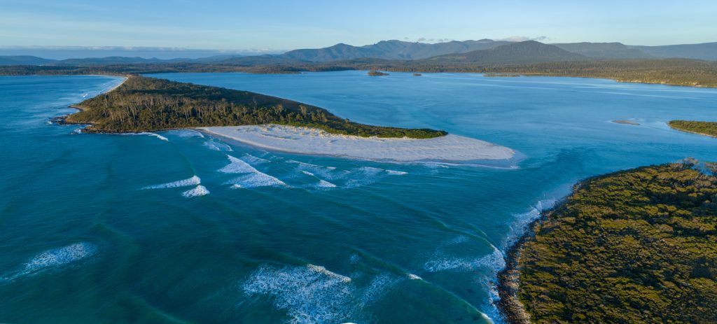 Aerial view of a coastal landscape with turquoise water, sandy beaches, lush green forests, and distant mountains under a clear sky. Waves gently break near the shore.
