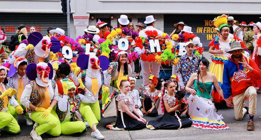 A vibrant group poses in colorful costumes, some holding large letters spelling COLOMBIA, during a festive street parade or celebration. Bright hats, ruffled skirts, and flower decorations fill the lively scene.