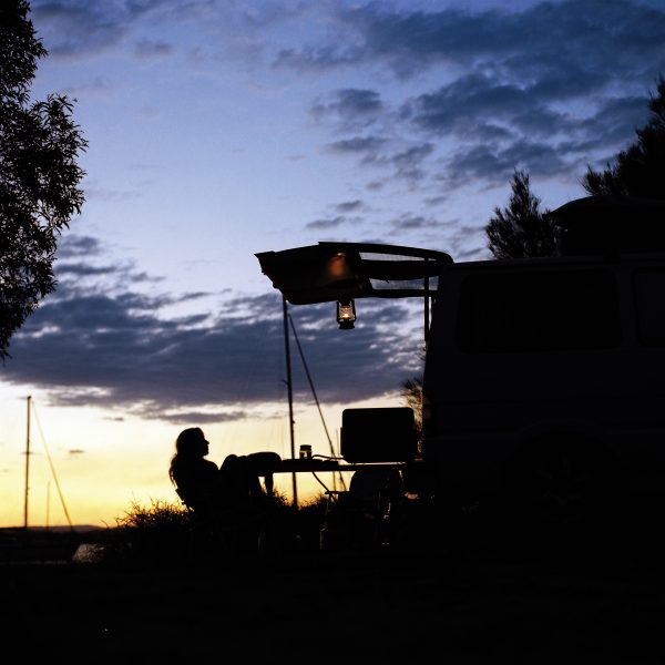 A person relaxes in a chair beside a van at sunset, with camping equipment set up under the van’s raised hatch and a lantern hanging above. Sailboats and trees are visible in the background against the evening sky.