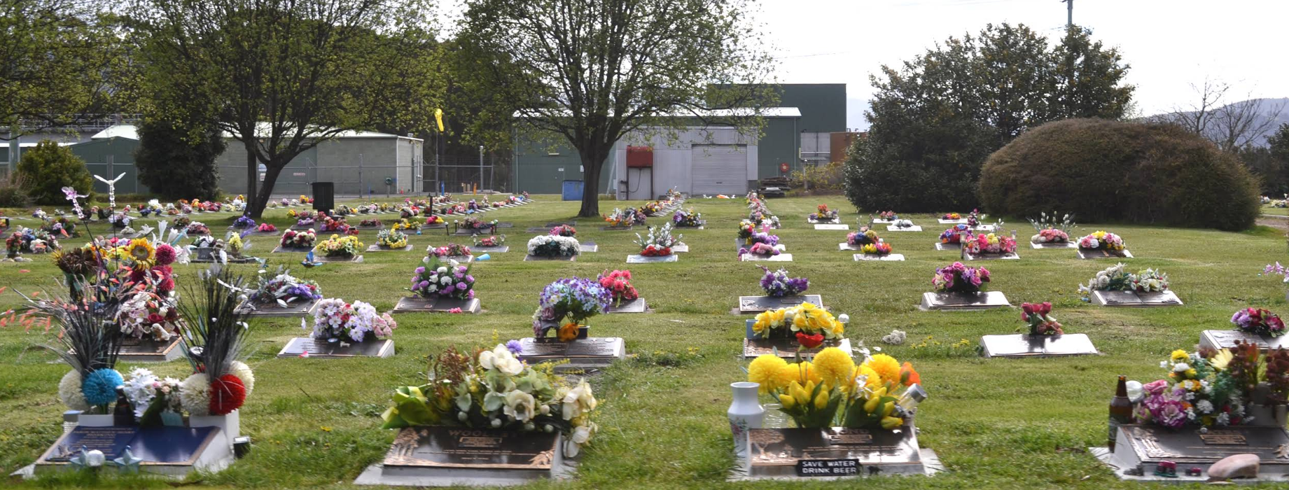 A cemetery with rows of graves adorned with colorful flower arrangements and plants. Green grass and several leafy trees are visible, along with buildings and a fence in the background.