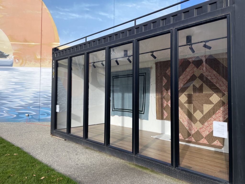 A glass-walled display space showcases two quilts on the walls. The structure is set on a sidewalk with a mural of a sunset over water painted on the adjacent building outside.