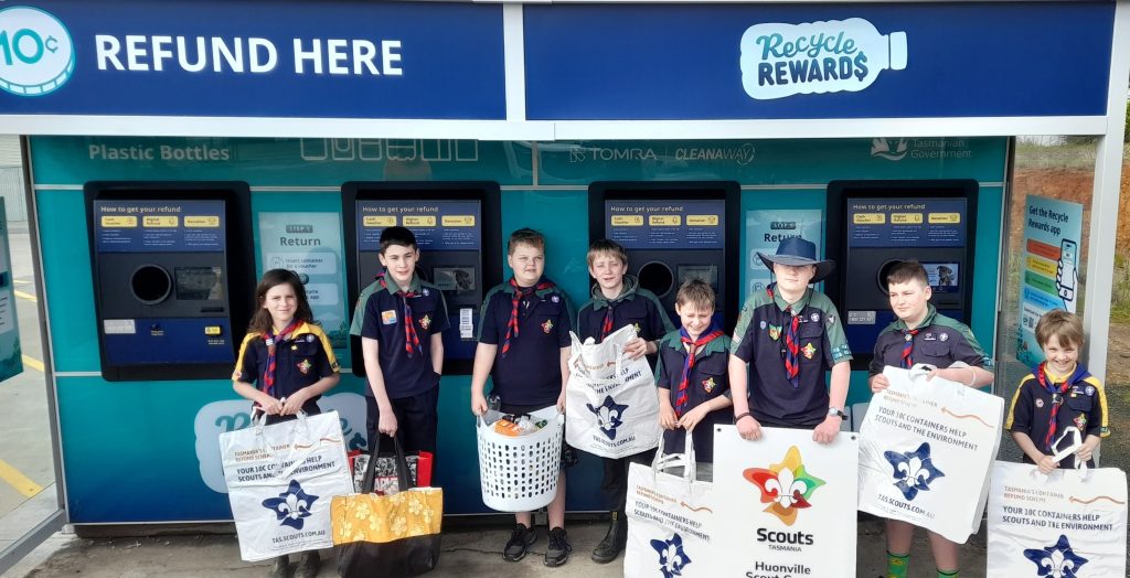 A group of children in Scouts uniforms stand in front of recycling machines holding bags and baskets filled with bottles for refund. A banner reads “Scouts Huonville” and the sign above the machines says “REFUND HERE” and “Recycle Rewards.”.