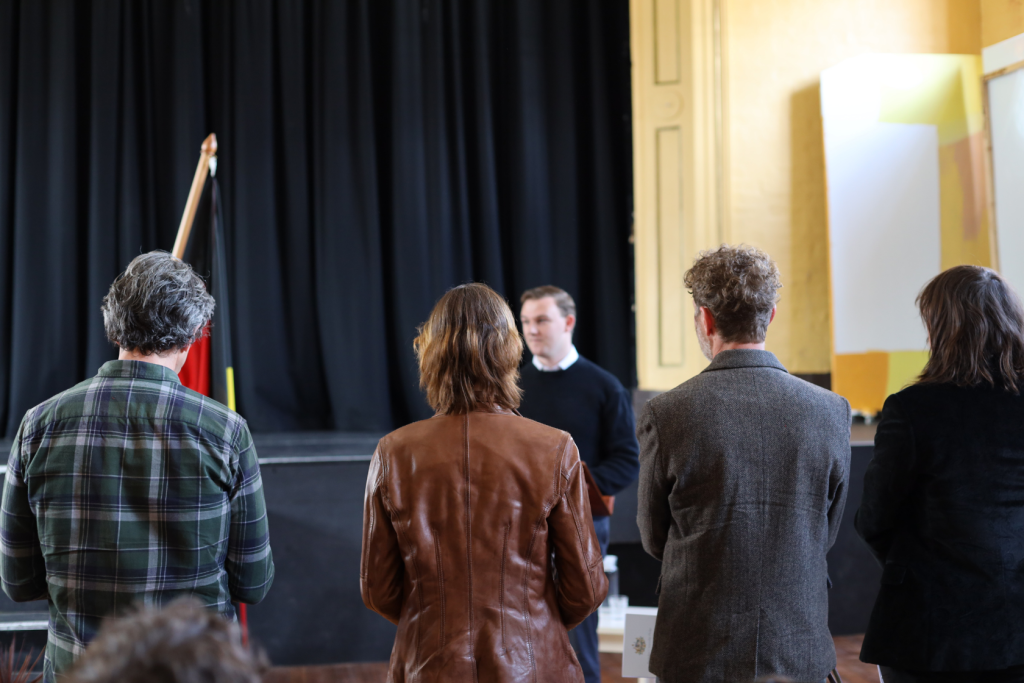 Four people seen from behind listen to a man speaking on a stage in front of a black curtain in a bright room. A flag is visible to the left, and the audience members wear jackets or coats.
