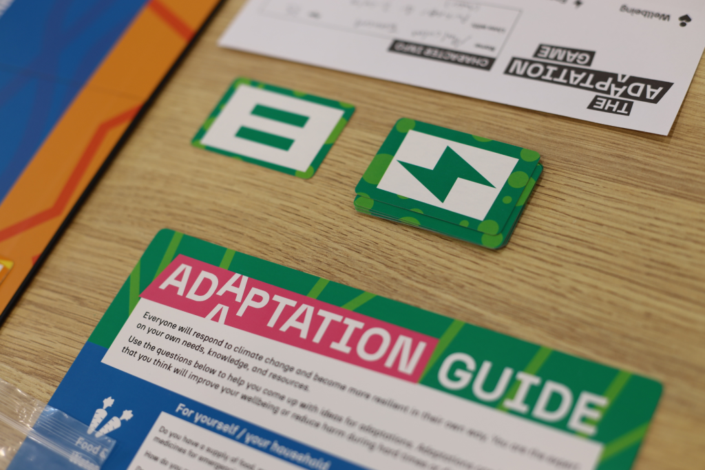 A close-up of a tabletop game called The Adaptation Game showing cards, including one with a green border and white symbol, and an ADAPTATION GUIDE instruction sheet with colorful headers.