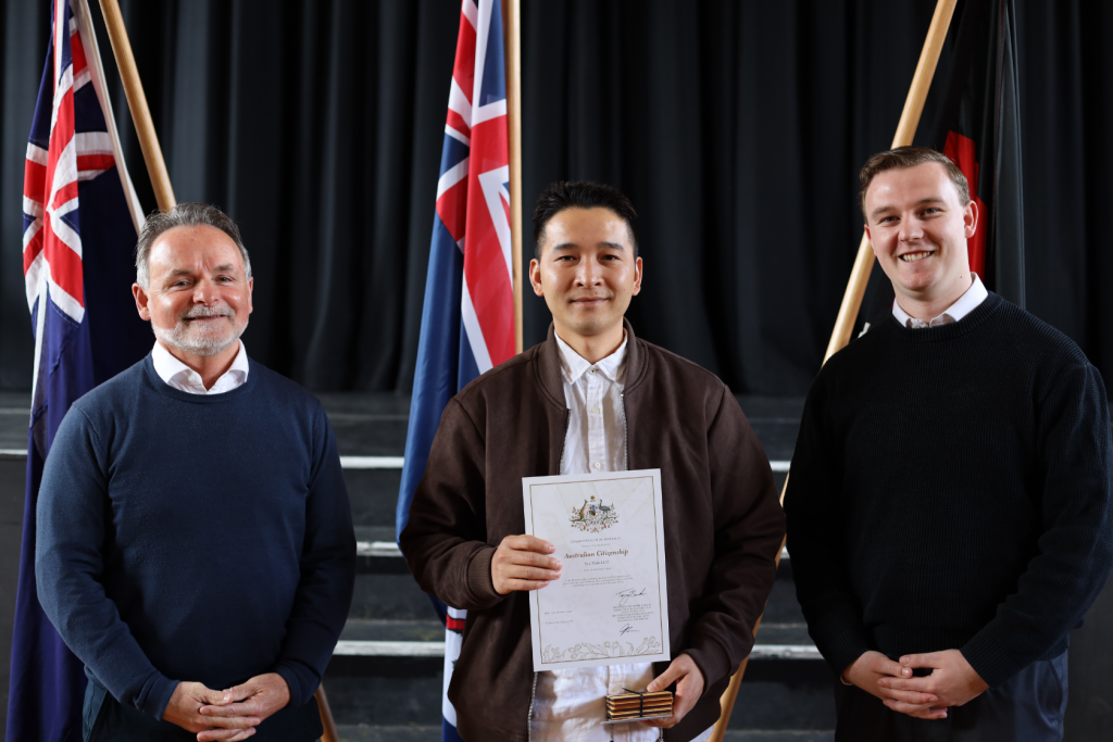 Three men stand in front of flags and black curtains. The man in the center holds a certificate and small box. All three are smiling and wearing sweaters or jackets. The scene appears to be a formal ceremony or award event.