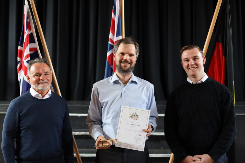 Three men stand side by side indoors in front of flags. The man in the center holds a certificate and a small wooden box, smiling, while the two others stand on either side, also smiling.