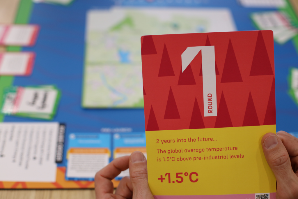 A person holds a game card reading Round 1: 2 years into the future... The global average temperature is 1.5°C above pre-industrial levels (+1.5°C). A colorful board game is blurred in the background.