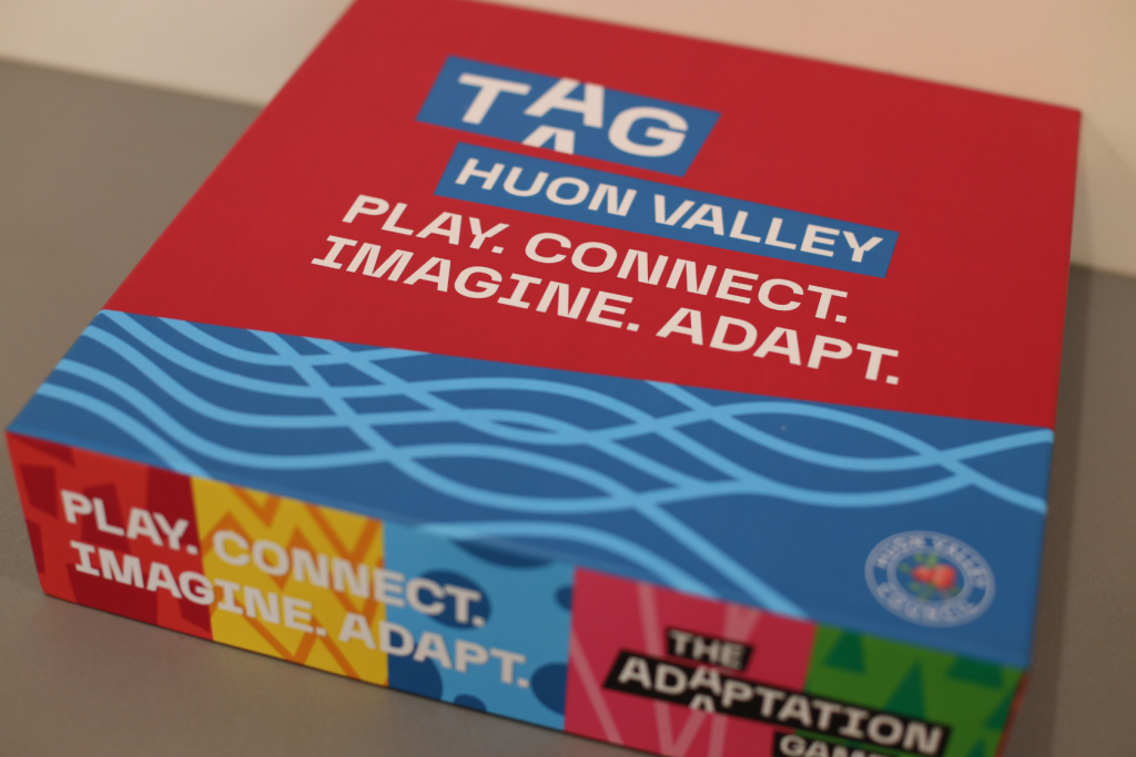 A red and blue game box titled TAG Huon Valley with the words Play. Connect. Imagine. Adapt. printed on the lid. The bottom edge shows colorful patterns and The Adaptation Game.