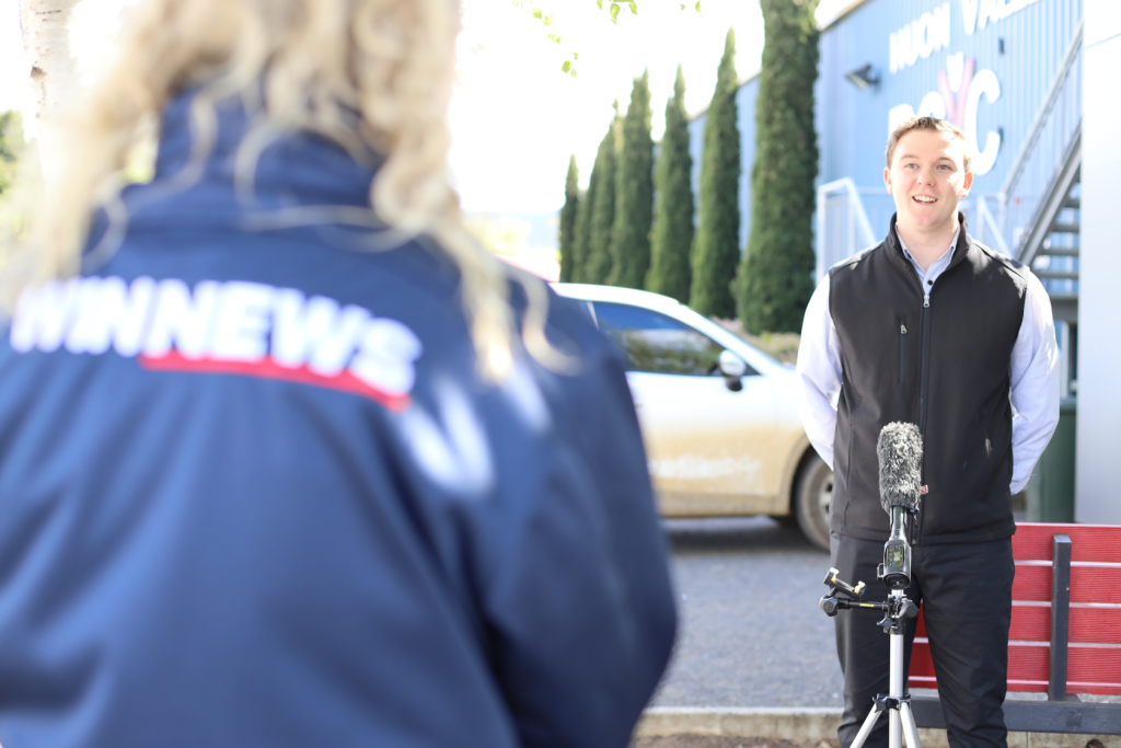 A man stands outdoors in front of a microphone on a tripod, being interviewed by a person wearing a blue WIN News jacket. A white car and trees are visible in the background.