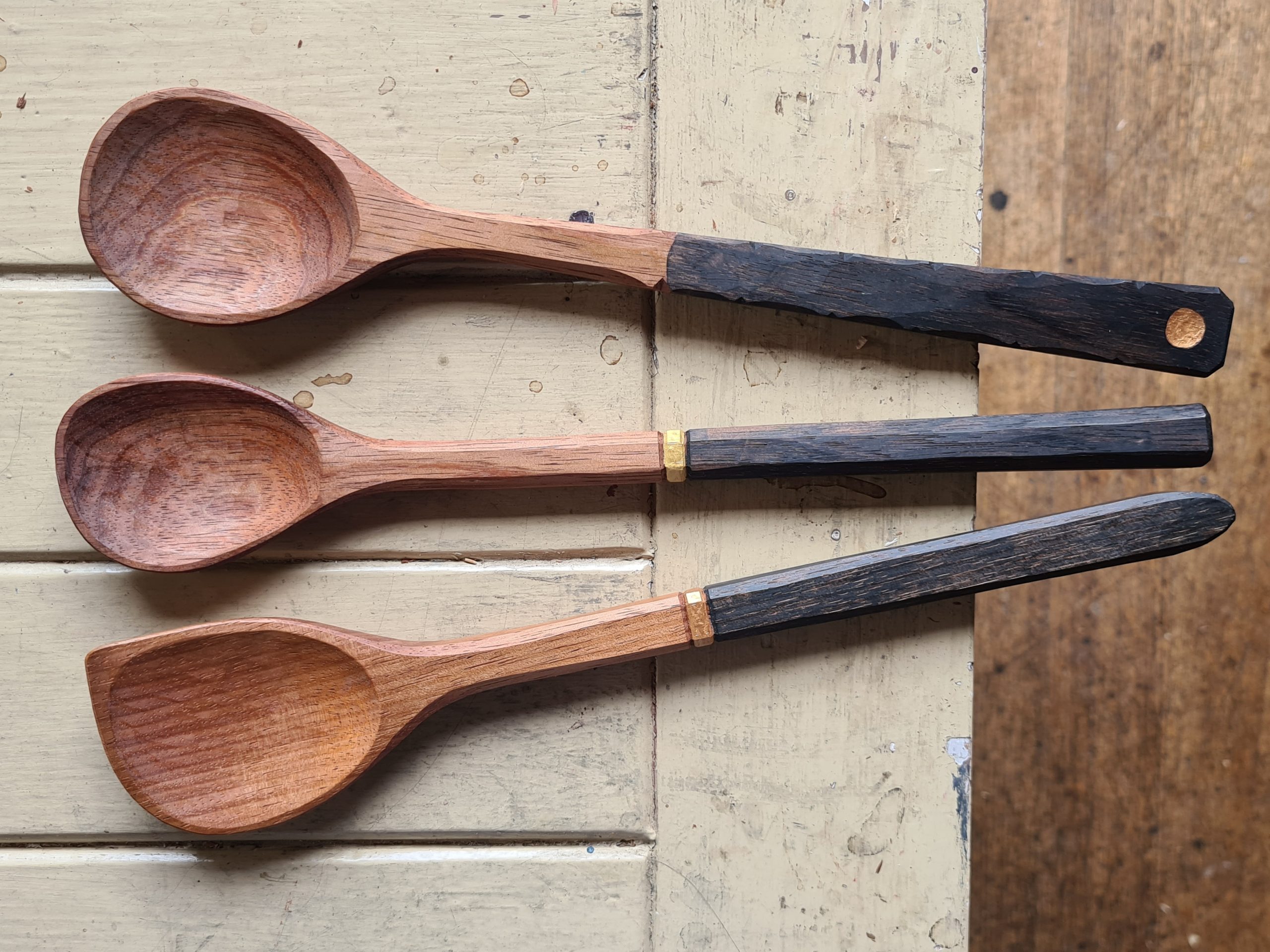 Three handcrafted wooden spoons with light brown bowls and dark, rustic handles are laid side by side on a worn, cream-colored wooden surface. The handles show distinct textures and one has a metal ring.
