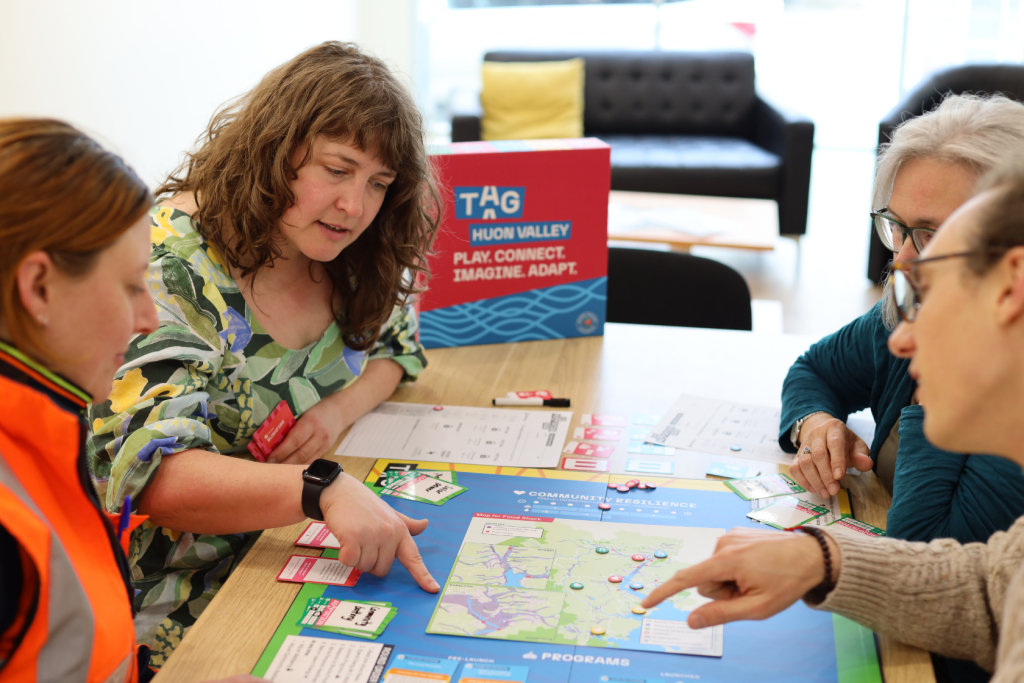 Four adults sit around a table playing a colorful board game. One woman points to a map on the board while others listen and engage, discussing strategies. Cards, papers, and game pieces are spread out on the table.