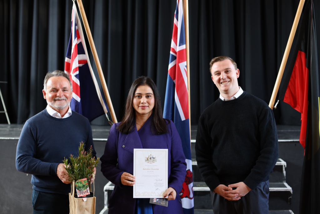Three people stand indoors in front of Australian and Aboriginal flags. The woman in the center holds a certificate, while the man on the left holds a plant in a bag. All are smiling at the camera.