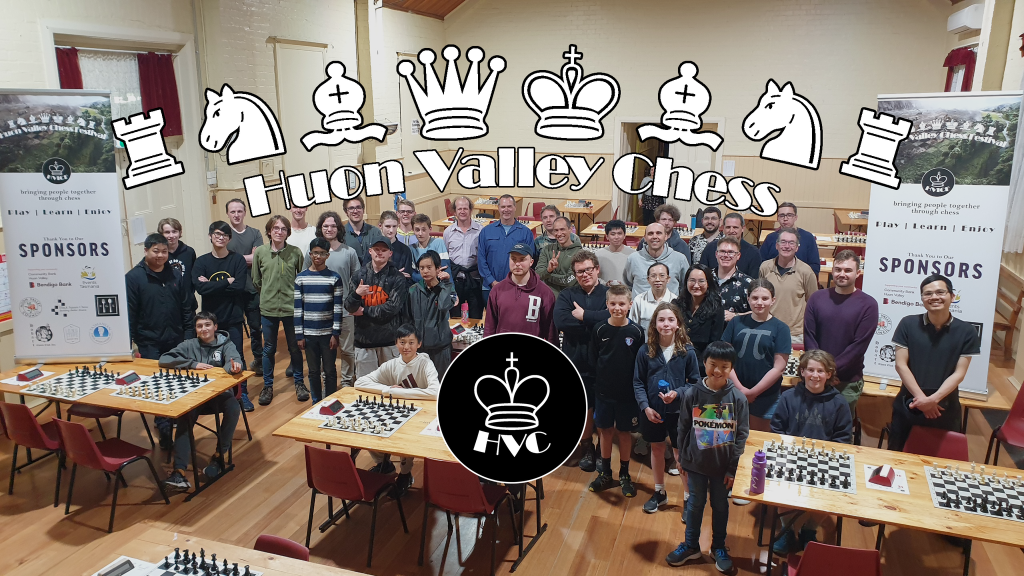 A large group of diverse people, including adults and children, pose in a hall with chess boards on tables. Banners and a Huron Valley Chess logo with chess piece icons are displayed above and in the center.