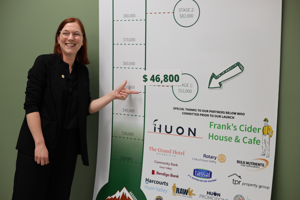 A smiling woman with red hair and glasses points to a fundraising chart showing $46,800, just below a $52,000 goal. Sponsor logos and acknowledgments are visible at the bottom of the chart.