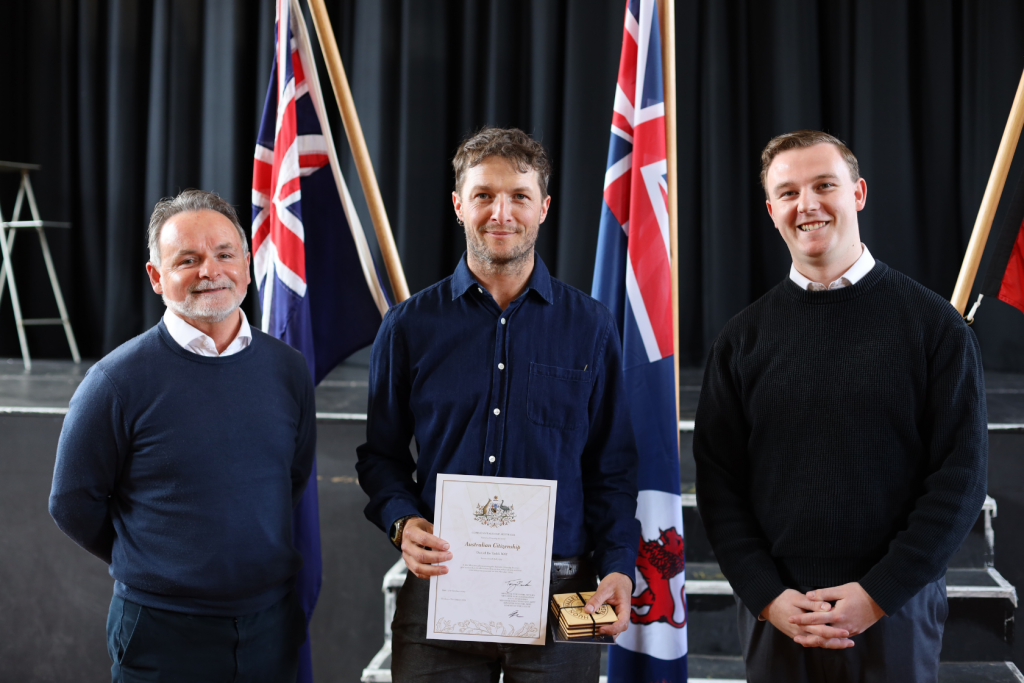 Three men stand indoors in front of two Australian flags. The man in the center holds a certificate and a small box, while the other two stand on either side, all smiling at the camera.