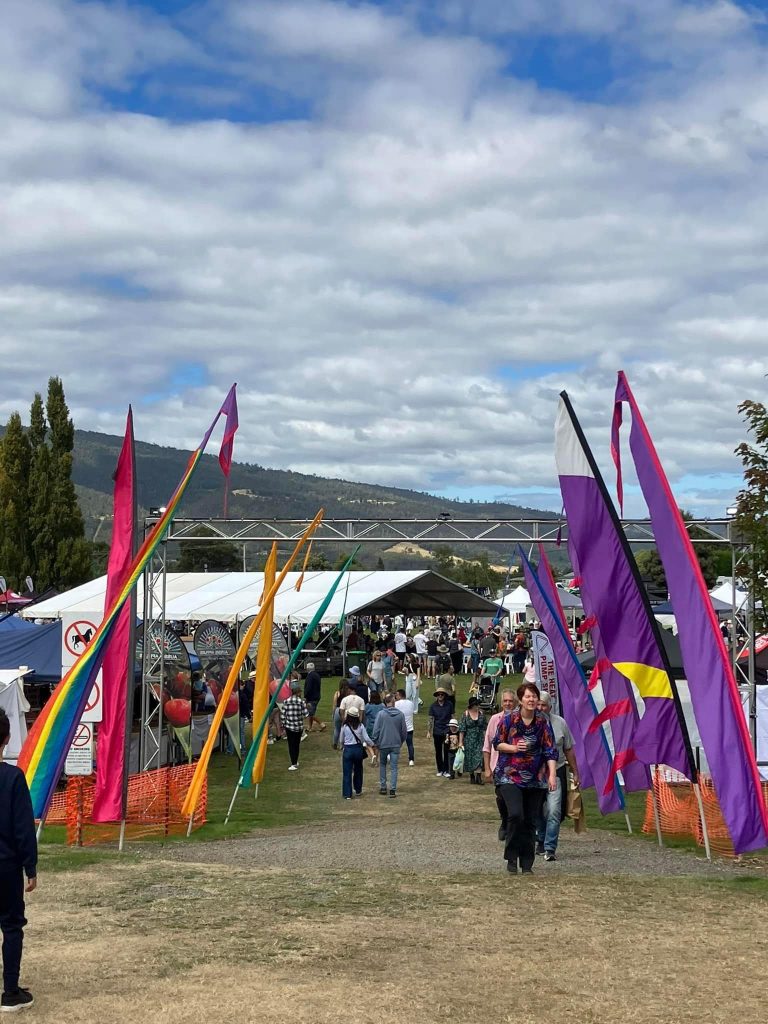People walk through colorful flag-lined paths at an outdoor festival with tents, mountains, and cloudy sky in the background. The event appears lively and festive under the daylight.