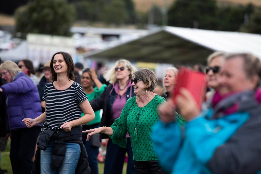 A group of adults, mostly women, are smiling and dancing outdoors at a daytime event. Some are wearing jackets and casual clothes, and one person in the foreground is taking a photo with a red device.