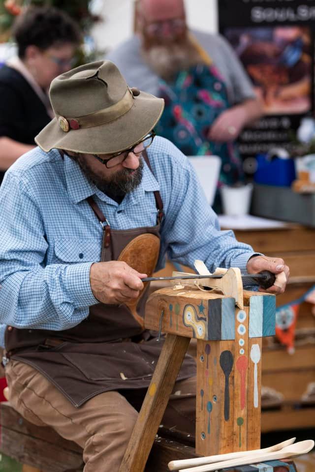 A man wearing glasses, a hat, and an apron carves wood with hand tools at a decorated workbench. People and colorful decorations are visible in the blurred background.