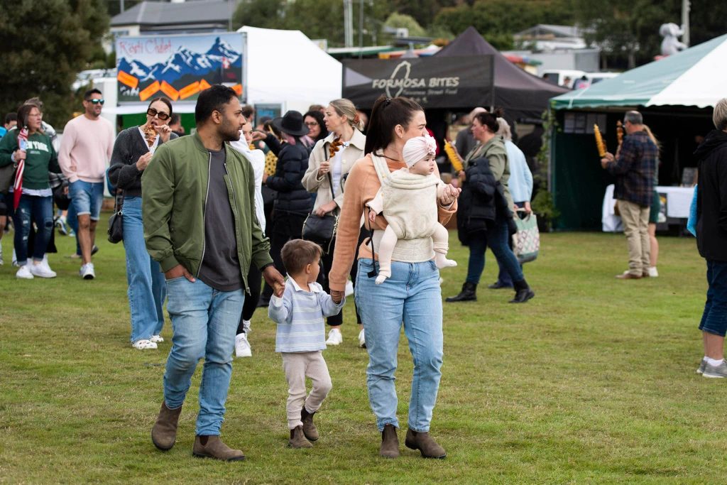 A family with two young children walks through an outdoor food festival. Other people are gathered around food stalls and tents in the background. The atmosphere is lively and casual.