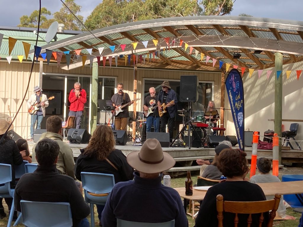 A band performs on an outdoor stage under a shelter decorated with colorful bunting, while an audience sits on chairs and benches, enjoying the live music under daylight.