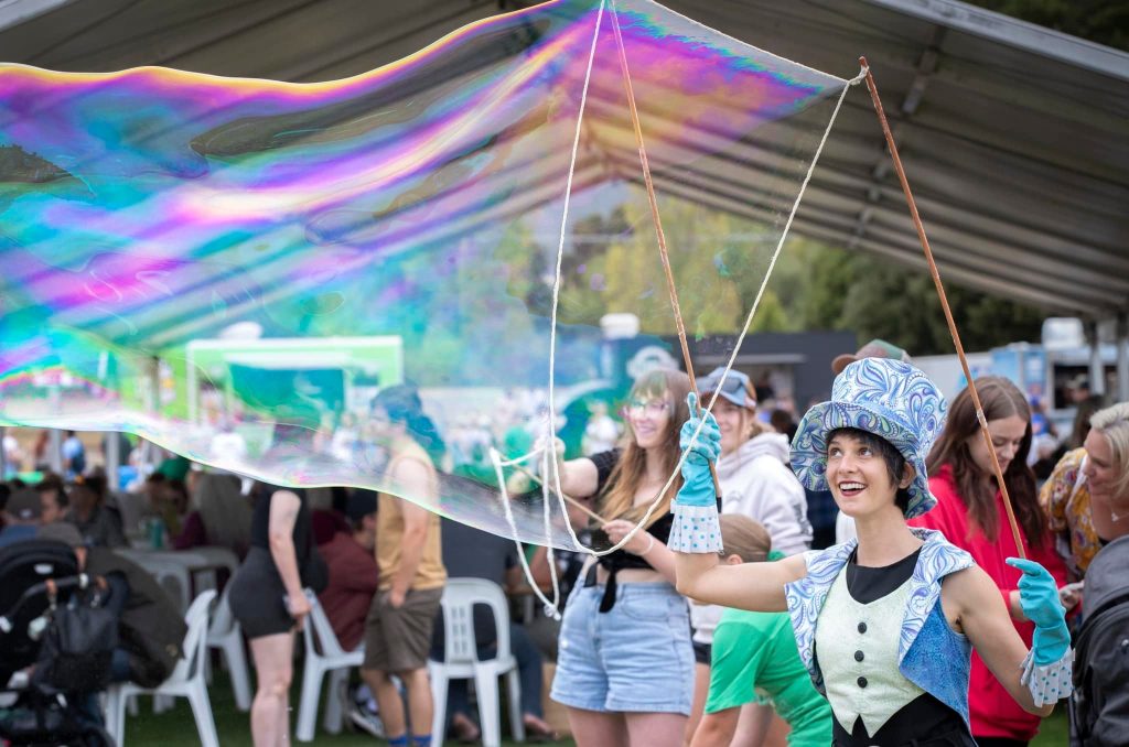 A woman in a colorful costume and patterned hat makes a large, iridescent bubble with sticks and string at an outdoor event under a canopy, while people watch and take photos in the background.