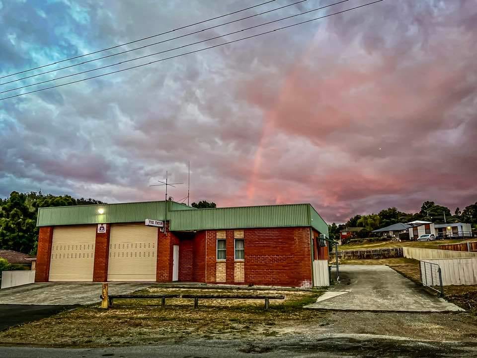 A small brick fire station with two garage doors sits under a dramatic, cloudy sky at sunset. A faint rainbow arcs above the building, and houses are visible in the background.