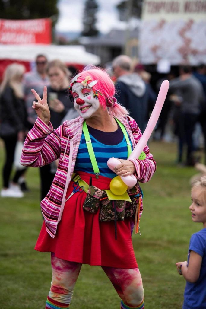 A smiling clown with pink hair, colorful makeup, a striped jacket, and a red skirt flashes a peace sign while holding balloon animals at an outdoor event. A small child stands nearby, and people are gathered in the background.