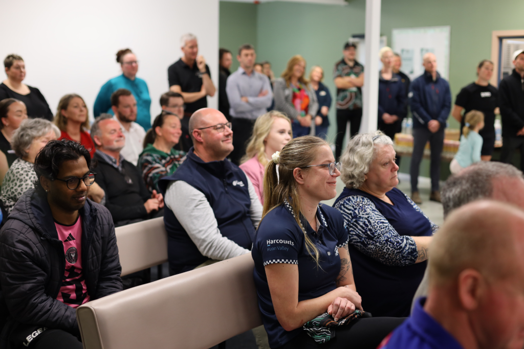 A diverse group of people sit attentively in rows of chairs at an indoor event, with some standing at the back. Most are facing forward, listening and engaged. The background includes green and white walls.