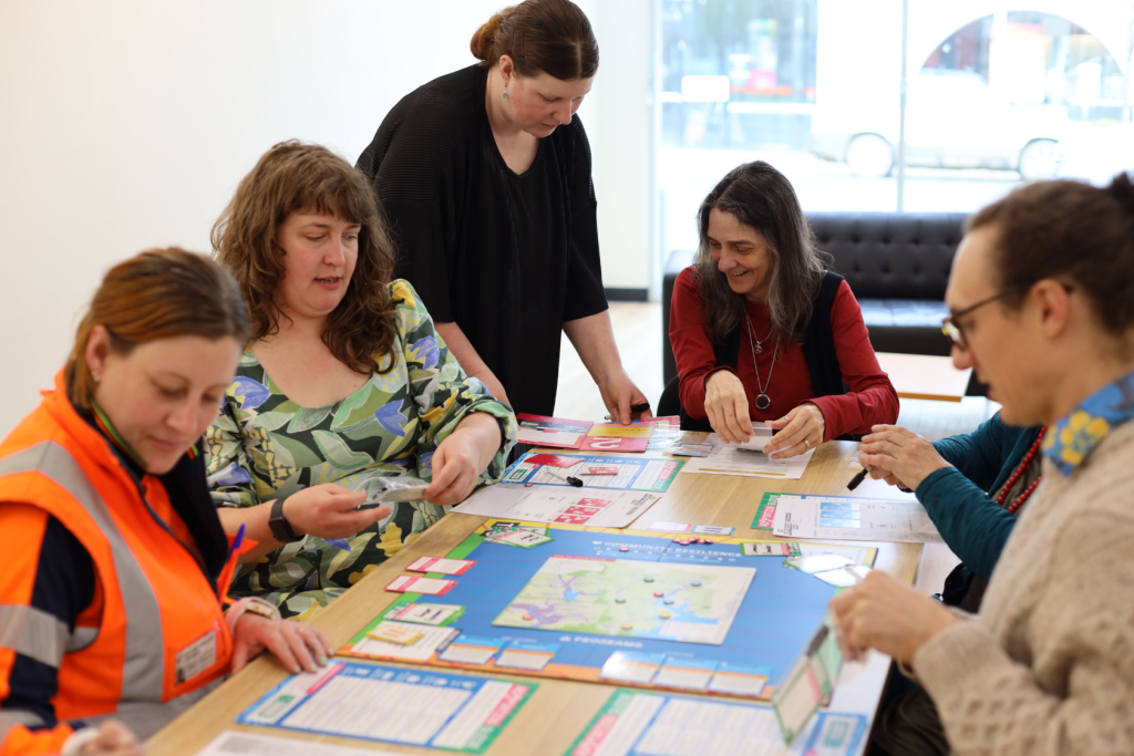 Five women are gathered around a table playing a board game with colorful cards and a map. One woman in an orange vest looks at cards, while others discuss and organize game pieces in a bright, modern room.