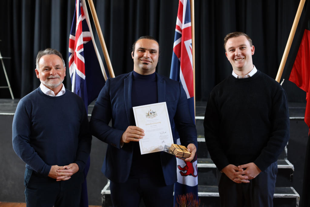 Three men stand indoors in front of Australian flags. The man in the center holds a certificate and a small box, while the other two stand on either side smiling. All are wearing dark sweaters.
